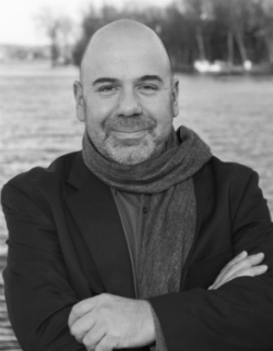 Black and white photo of Ivan Jenson standing in front of a lake, wearing a scarf and smiling with his arms crossed